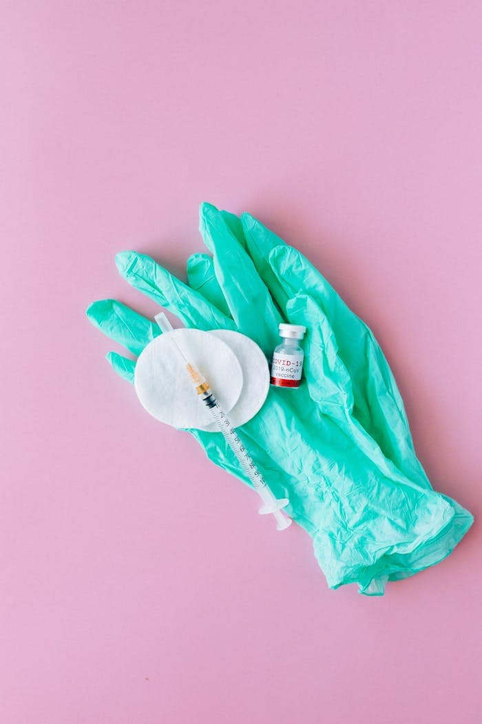 Flat lay of medical gloves, syringe, and vial on pink background.